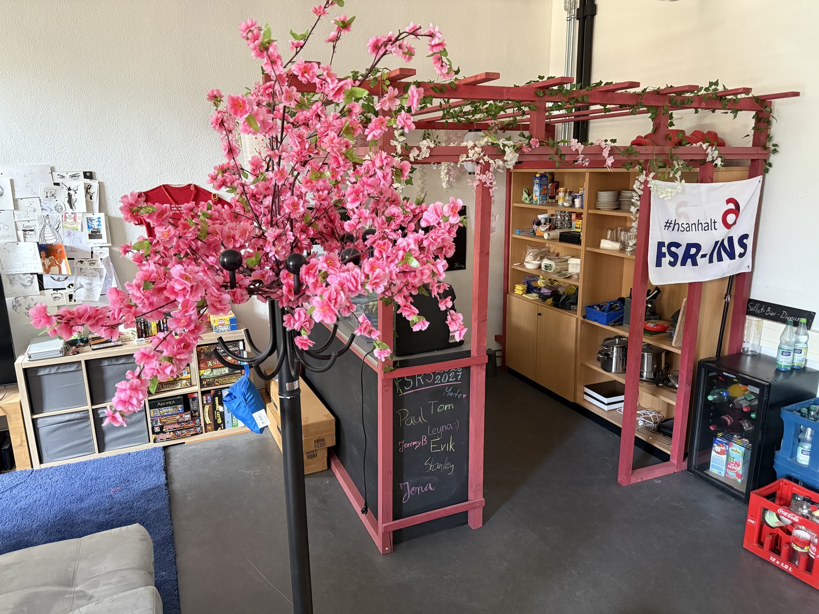 Blick in das StudiLink mit Sitzbereich, Brettspielen, Küchenregalen und rosa Blütendekoration